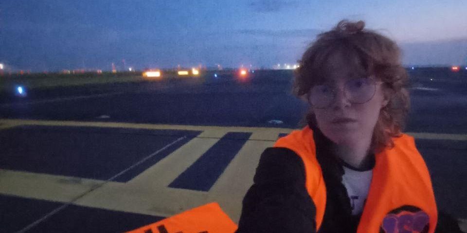 Person mit orangefarbener Weste hält ein Schild mit der Aufschrift 'OIL KILLS' auf einem Flughafen in Berlin bei Sonnenaufgang