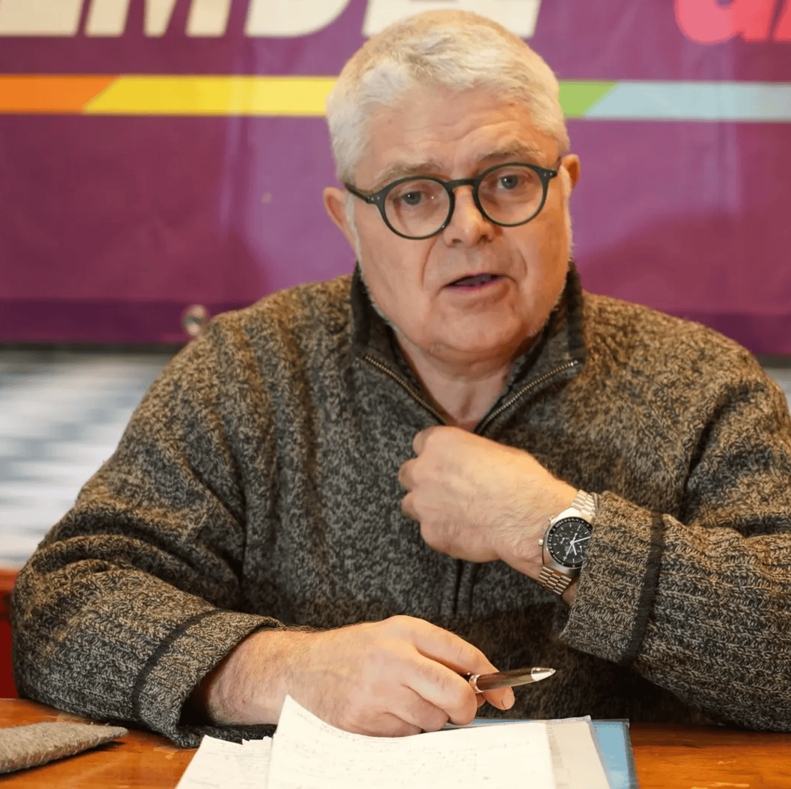 Homme âgé avec des lunettes et un pull en laine grise, assis à une table avec des papiers et un stylo.
