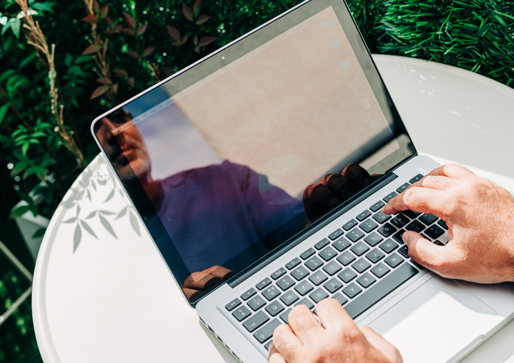 Mann arbeitet im Freien an einem Laptop, der in der Sonne auf einem weissen Tisch steht, umgeben von grünem Laub.