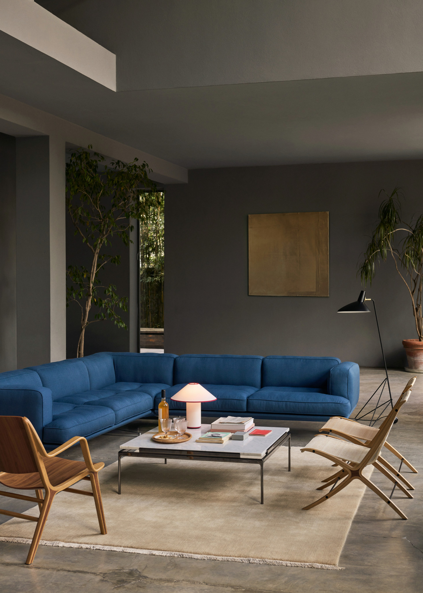 Modernes Wohnzimmer mit blauem Ecksofa, Couchtisch mit Lampe, Holzstühlen und Zimmerpflanzen vor grauen Wänden.