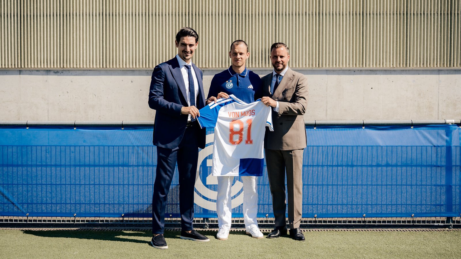 Drei Männer präsentieren ein blau-weisses Fussballtrikot mit der Nummer 81 vor einem Fussballplatz.