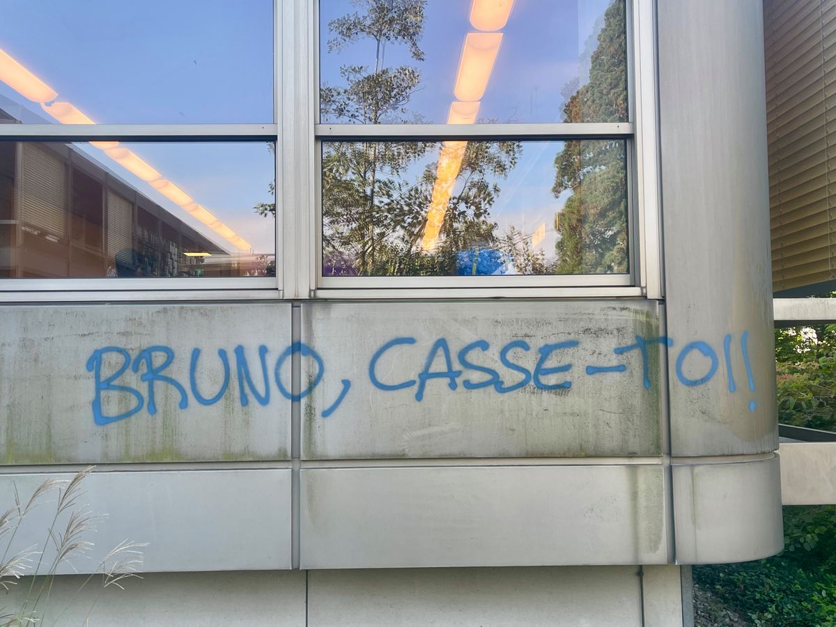 Graffiti bleu sur un mur, lisant 'BRUNO, CASSE-TOI!!' sous une fenêtre réfléchissant des arbres et des lumières de bâtiment.