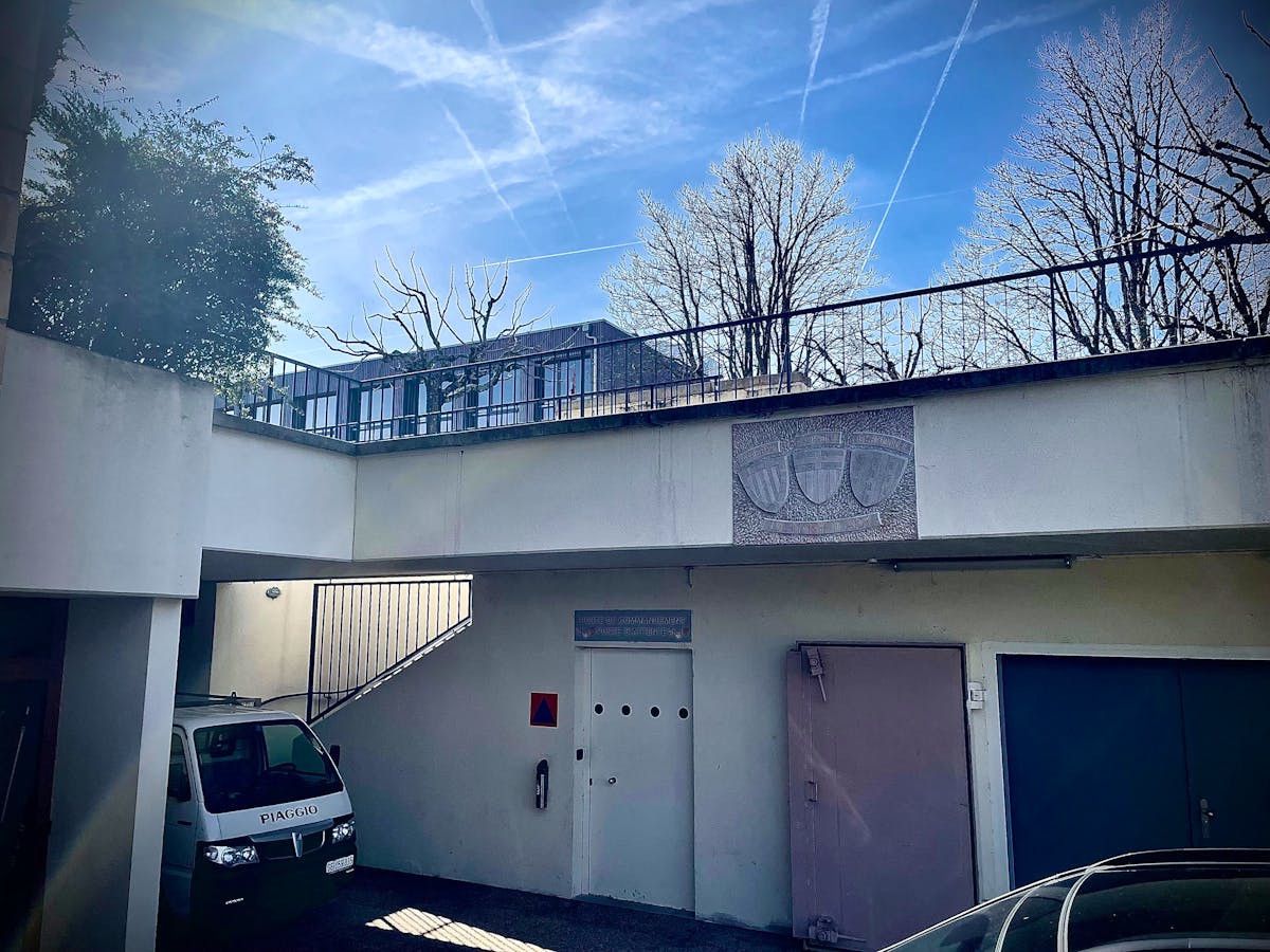 Entrée d'un bâtiment avec un van blanc stationné à gauche, un pont au-dessus avec une gravure sur le mur, et un ciel bleu avec des arbres sans feuilles en arrière-plan.