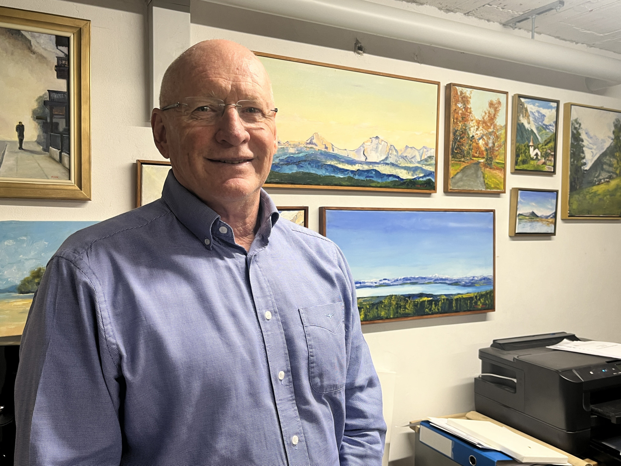 Mark Nolan steht lächelnd vor einer Wand mit mehreren Landschaftsgemälden, darunter Schweizer Bergpanoramen im Stil Ferdinand Hodlers. Mark Nolan steht lächelnd vor einer Wand mit mehreren Landschaftsgemälden, darunter Schweizer Bergpanoramen im Stil Ferdinand Hodlers.