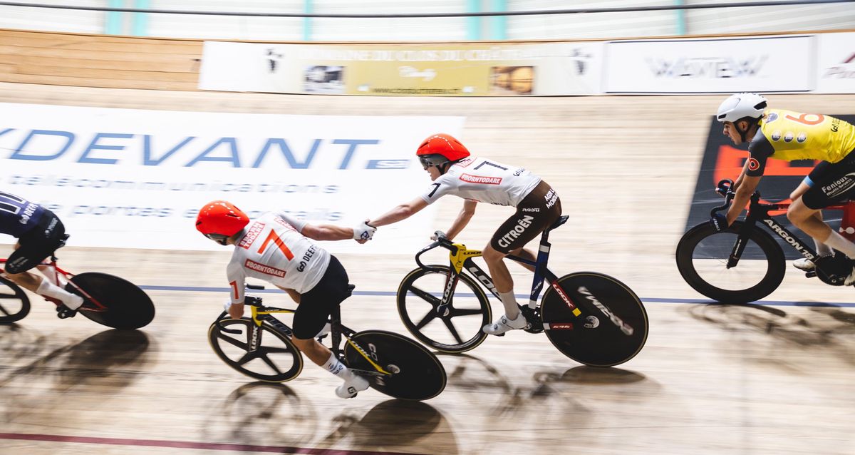 Deux cyclistes portant des casques rouges s’échangent un passage de relais sur une piste de vélodrome, suivis de près par un troisième cycliste.