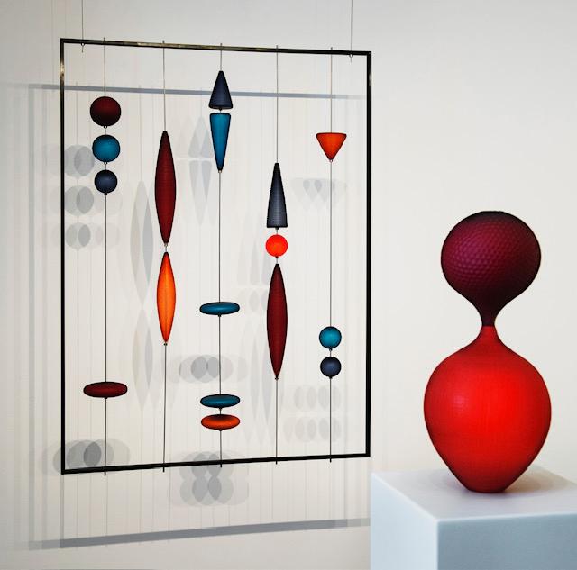 Installation artistique moderne avec des figures géométriques en suspension à gauche et une sculpture rouge abstraite sur un socle à droite.