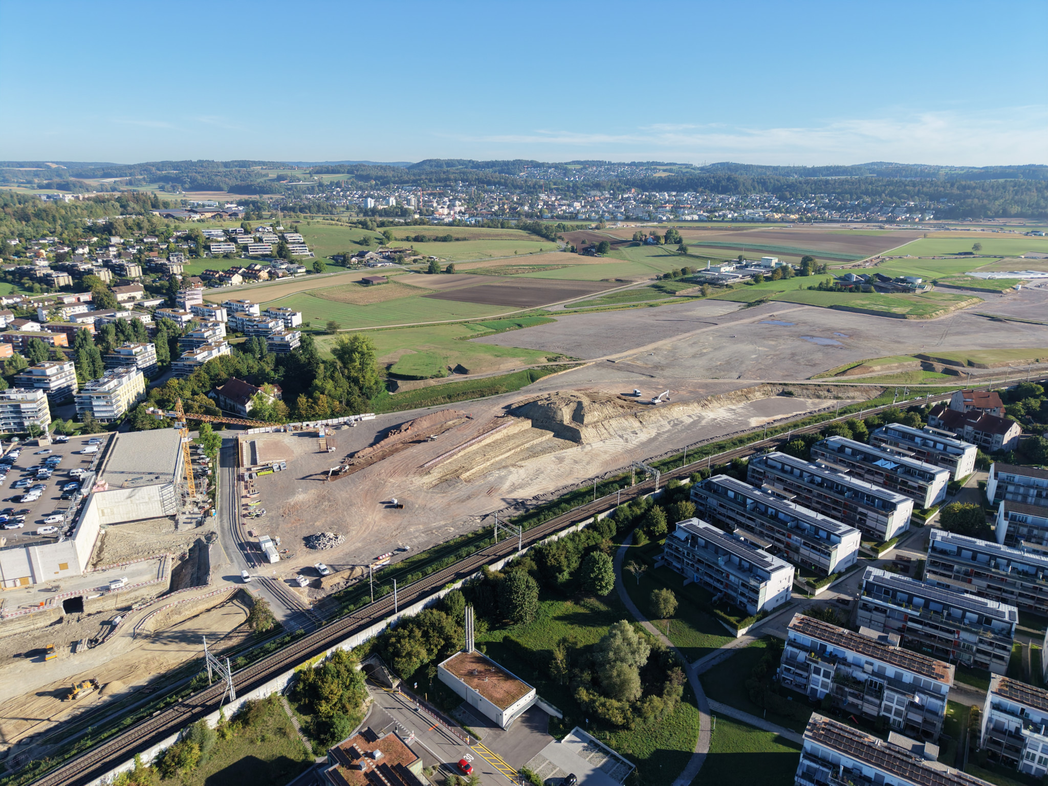 Luftaufnahme einer Baustelle neben Wohngebäuden und einer Bahnlinie, mit Feldern und einer Stadt im Hintergrund.