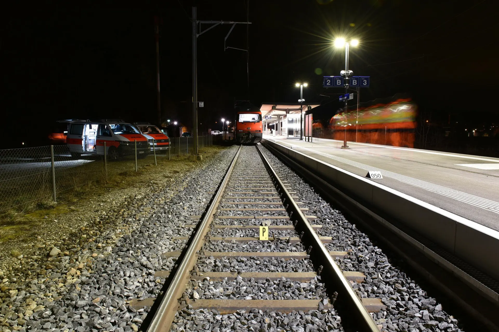 Nächtliche Bahnhofsszene mit Gleisen, stehendem Zug auf Perron und Einsatzfahrzeugen neben dem Gleis, gelbes Spurensicherungsschild «P» auf den Schienen.