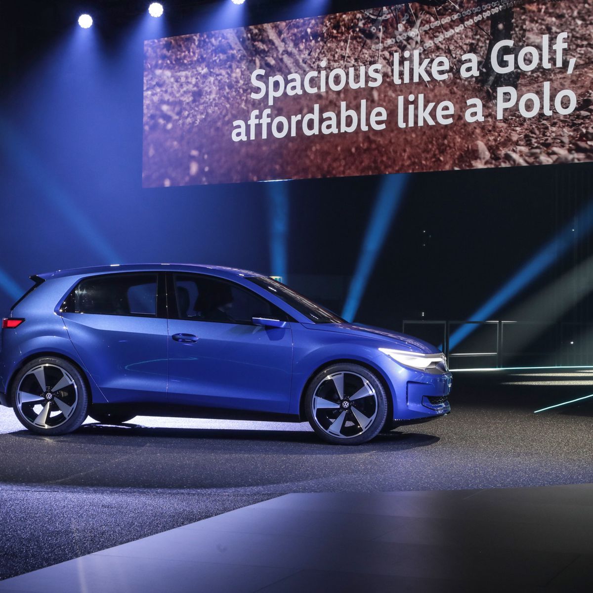 Der VW ID.2 unter Scheinwerferlicht, präsentiert mit dem Slogan ’Spacious like a Golf, affordable like a Polo’ im Hintergrund.