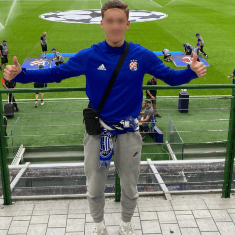Un jeune supporter portant un sweat-shirt bleu avec le logo de l’équipe, une écharpe et un jogging gris, pose avec deux pouces levés devant un terrain de football avec le logo de la Ligue des Champions sur le gazon.