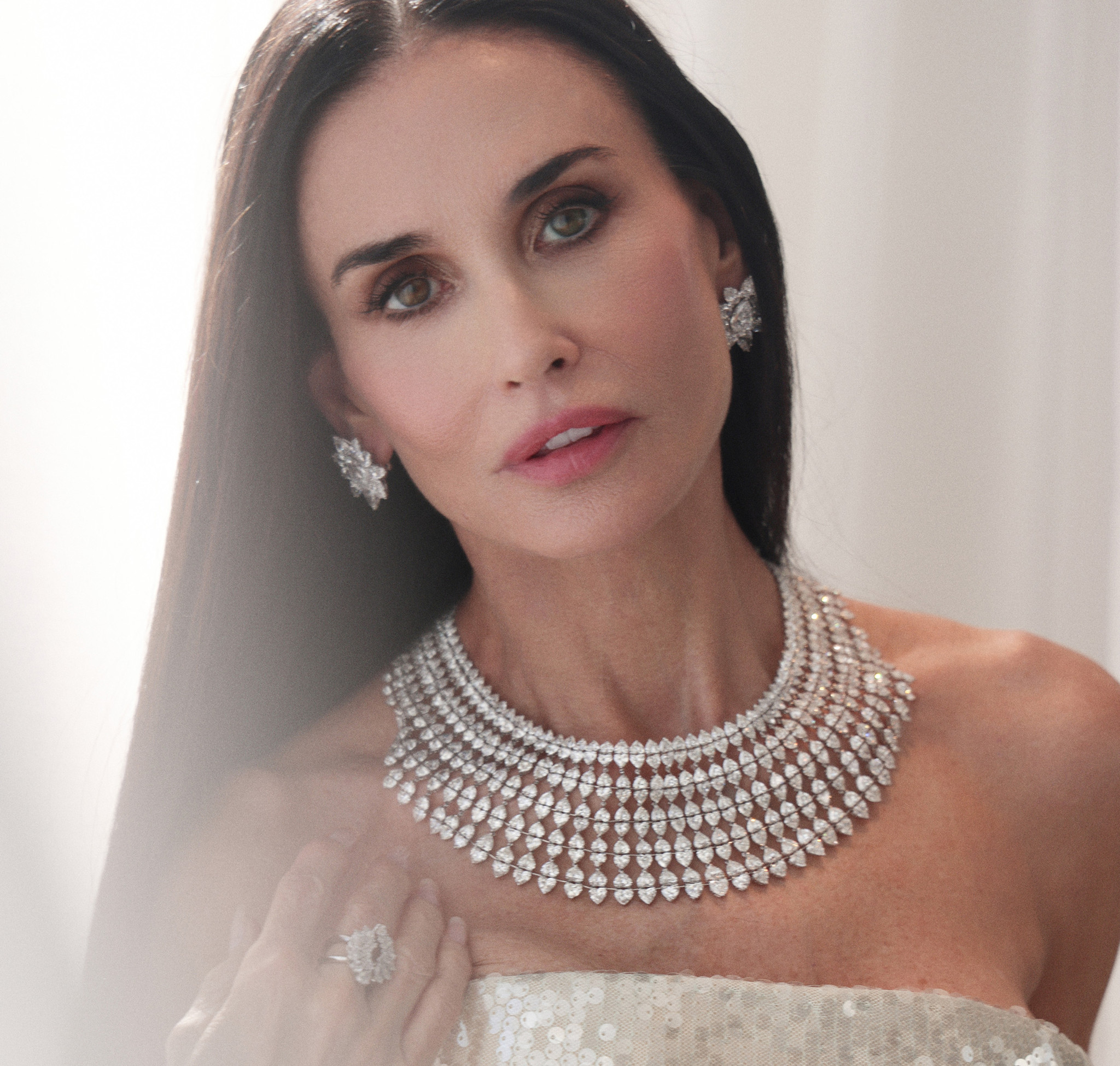 A dark-haired woman wearing a lavish diamond necklace, matching earrings and an elegant white dress.