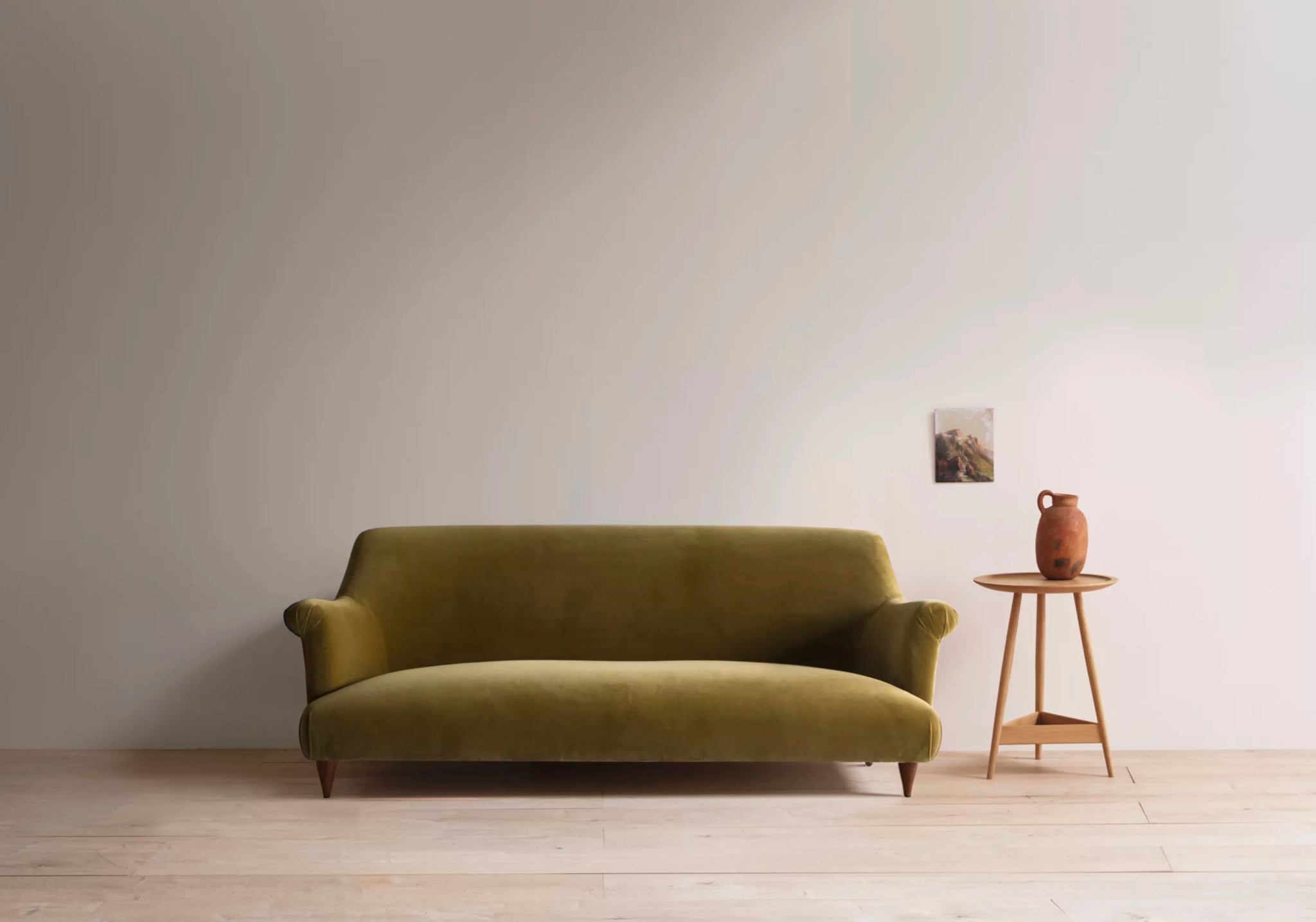 Olivgrünes Samtsofa mit Holzbeinen neben einem runden Beistelltisch mit Terrakotta-Vase vor heller Wand.