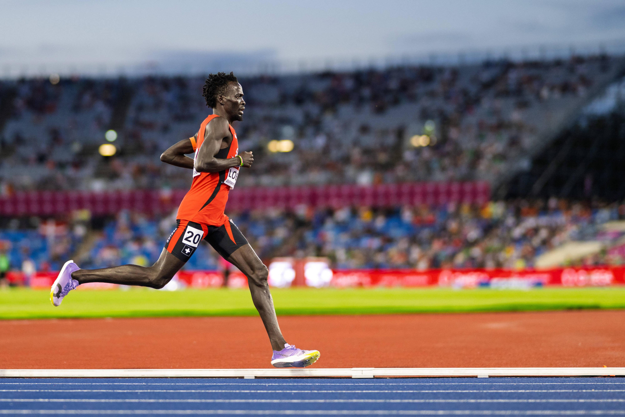 Läufer mit orangem Trikot und Startnummer 20 sprintet auf blauer Tartanbahn in einem Leichtathletikstadion.