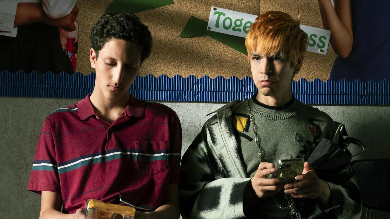 Deux jeunes hommes assis côte à côte, l’un portant un polo rouge et l’autre vêtu d’un pull gris avec une coiffure blonde. Ils tiennent des objets dans leurs mains, assis devant un tableau d’affichage.