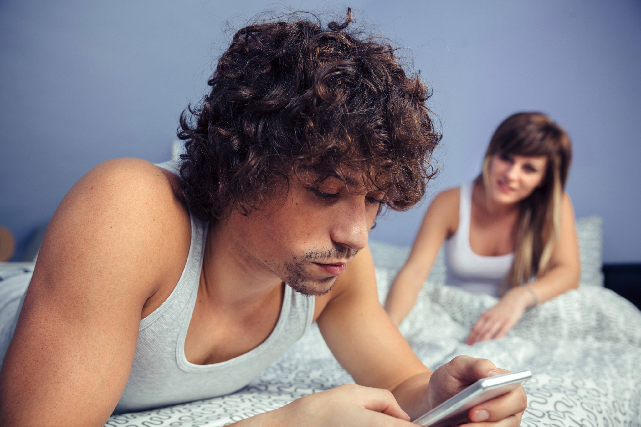 Un homme allongé sur un lit, concentré sur son smartphone, tandis que sa partenaire le regarde en arrière-plan, illustrant l’absence dans le couple.