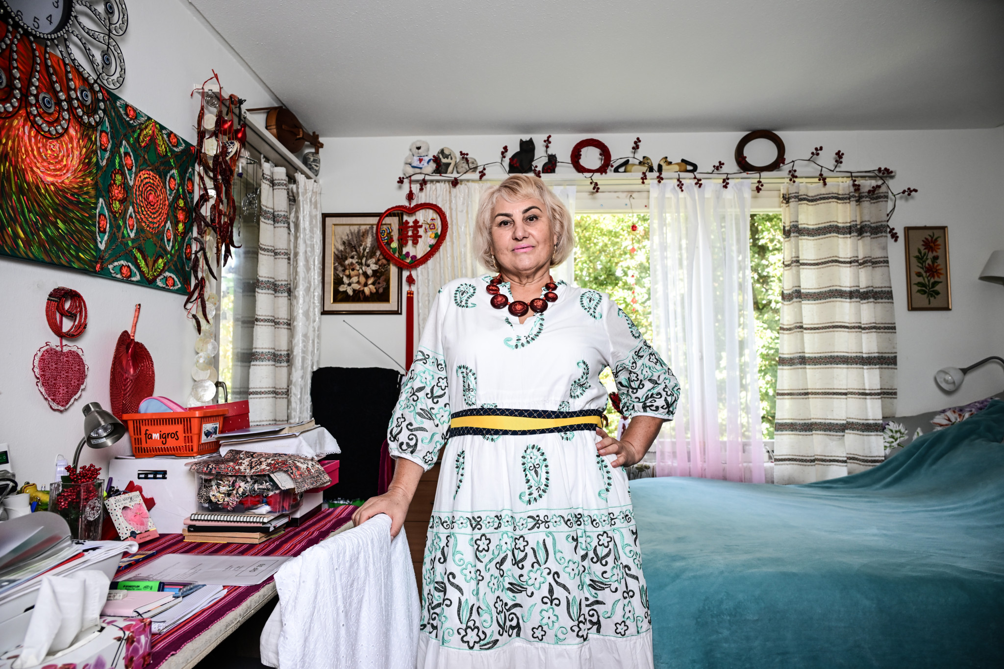 Valentyna Kleiner, 60-jährige Ukrainerin, steht in ihrem schlicht eingerichteten Zimmer in einem gemusterten Kleid mit rotem Schmuck.