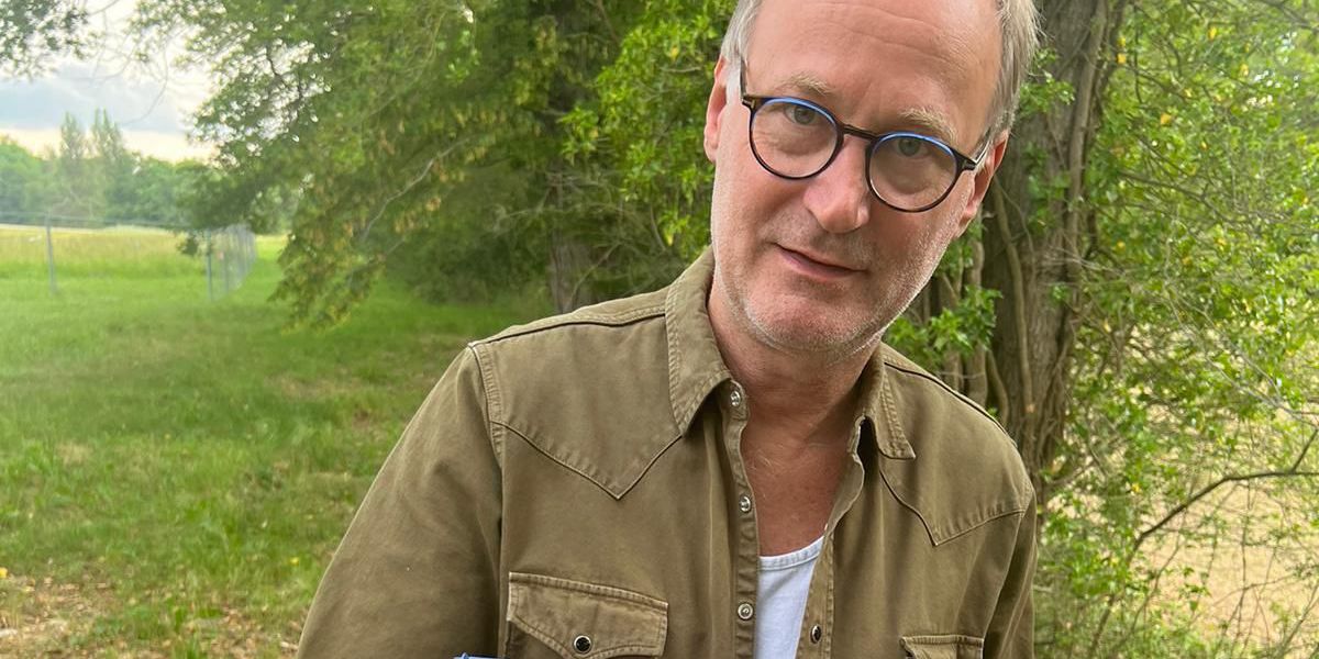 Un homme portant des lunettes et une chemise verte tient un panier rempli de champignons variés, debout dans un environnement naturel.