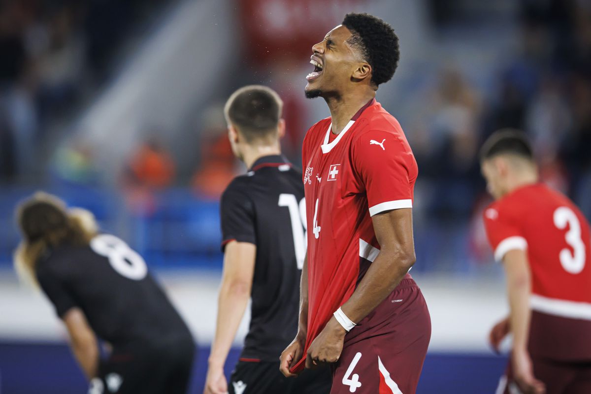 Un joueur de football suisse, vêtu d'un maillot rouge avec le numéro 4, célèbre sur le terrain tandis que des adversaires en noir sont vus en arrière-plan.