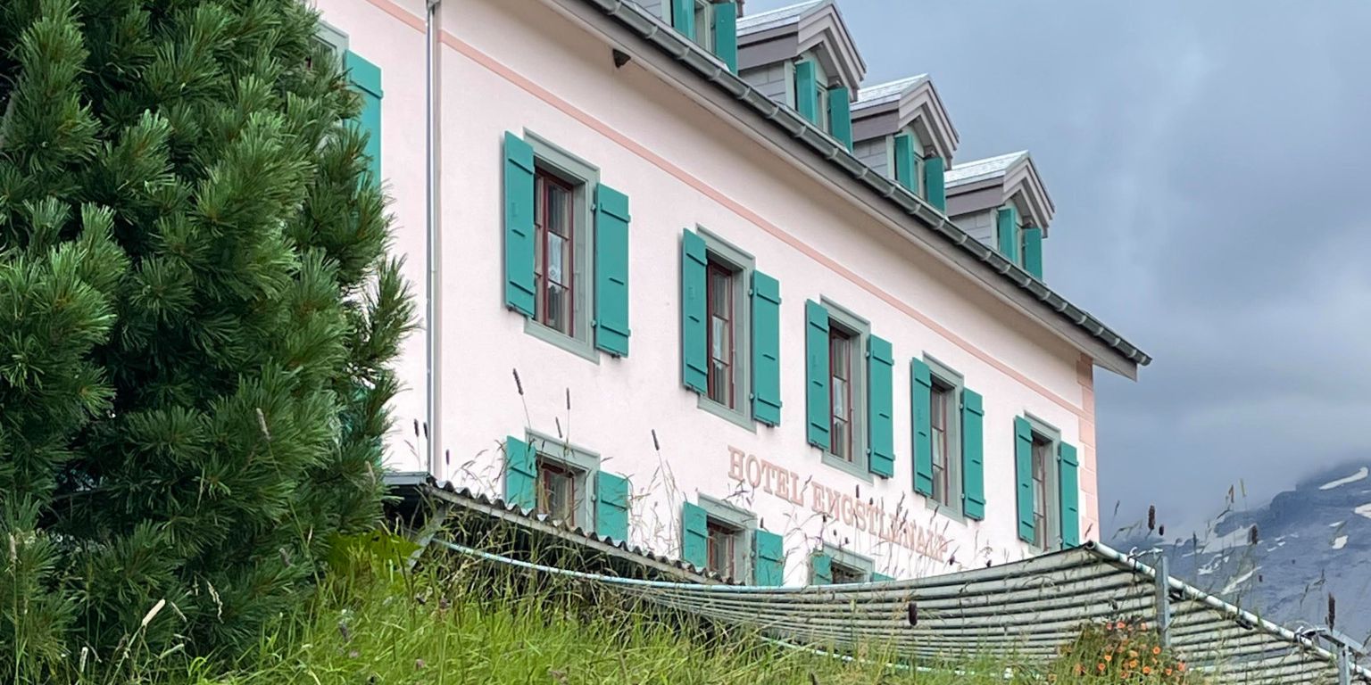 Une maison rose à plusieurs étages avec des volets verts, entourée de verdure et d'herbe, avec des montagnes en arrière-plan sous un ciel nuageux.