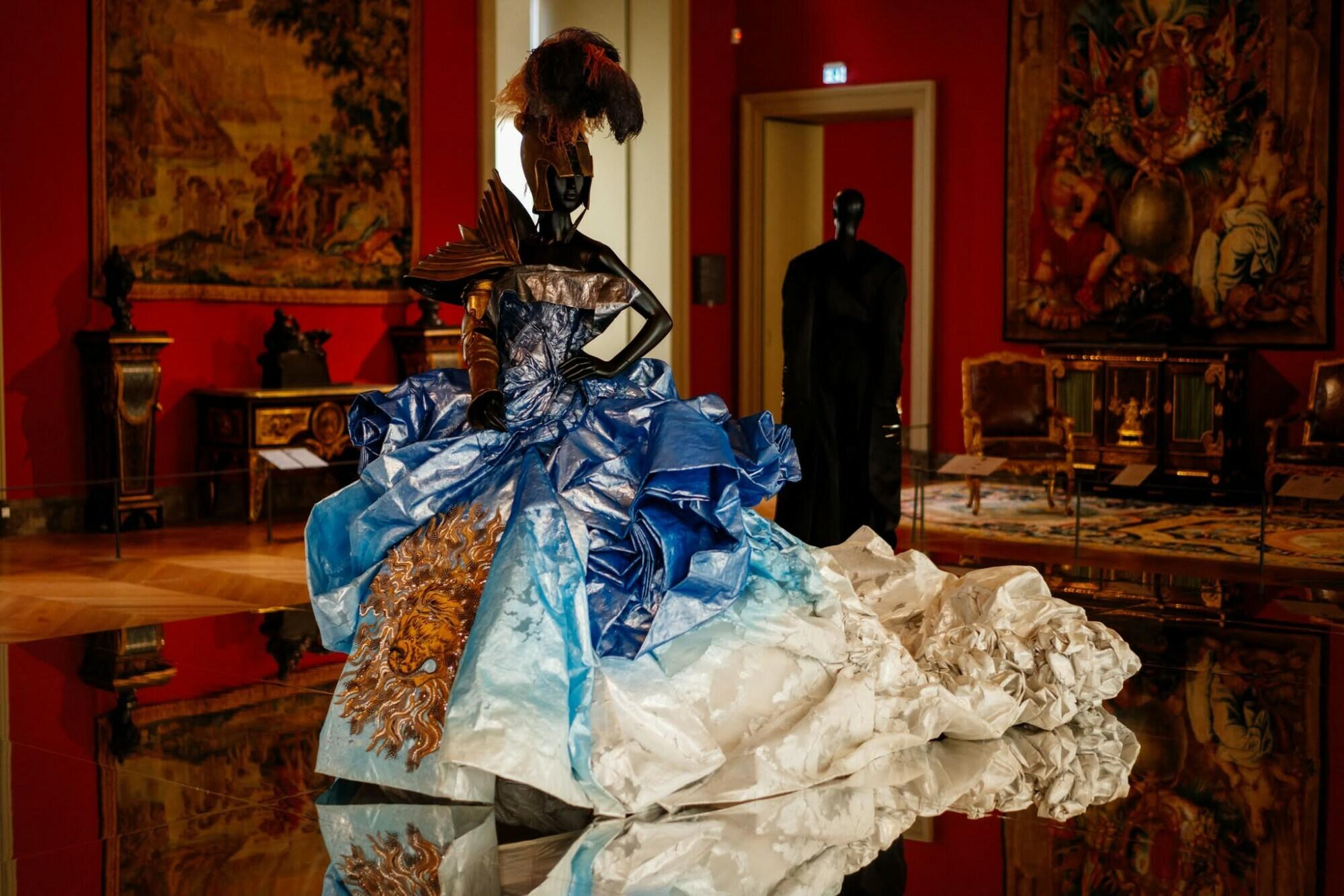 Robe de haute couture bleu et blanc ornée de plumes dans une galerie d’art au décor riche.