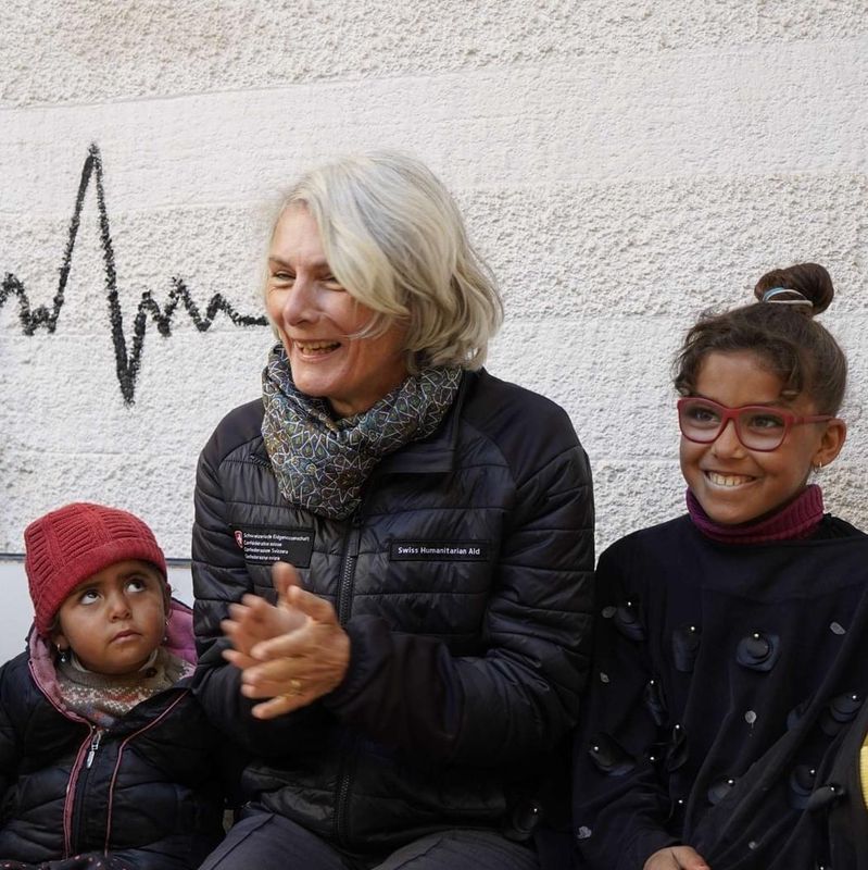 Gruppe von lächelnden Menschen, darunter zwei Erwachsene und drei Kinder, sitzt nebeneinander auf einer Bank vor einer bemalten Wand.