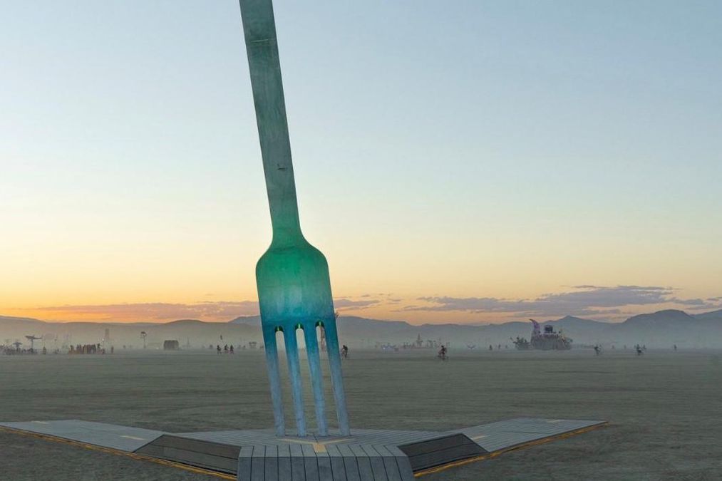 Grande fourchette dressée dans un paysage désertique au coucher du soleil, sur un socle représentant une route.