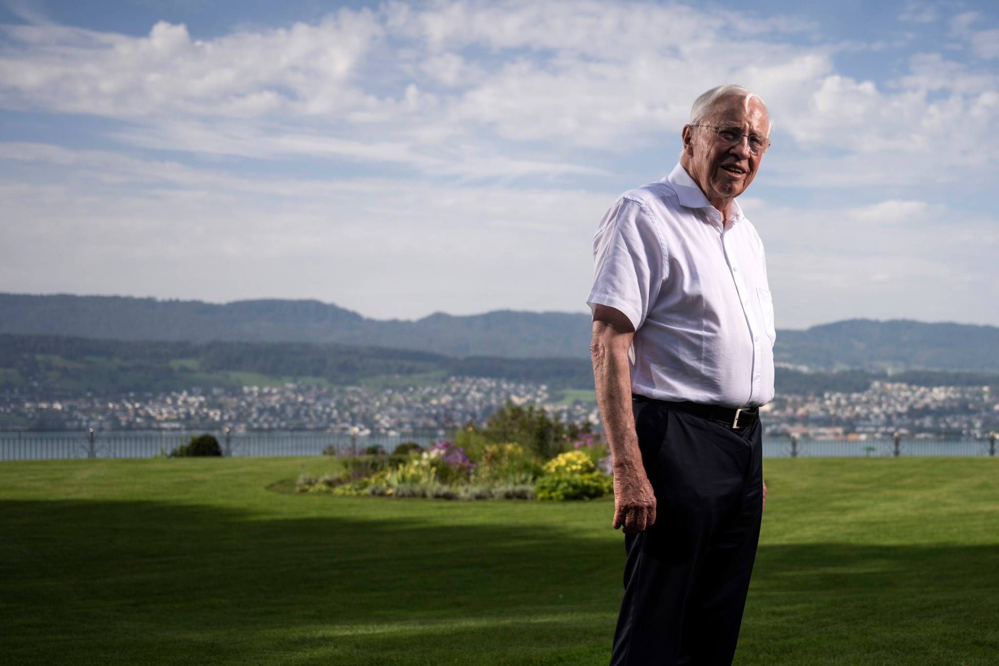 Christoph Blocher steht lächelnd auf einer grünen Wiese, im Hintergrund der Zürichsee und Hügel unter blauem Himmel.