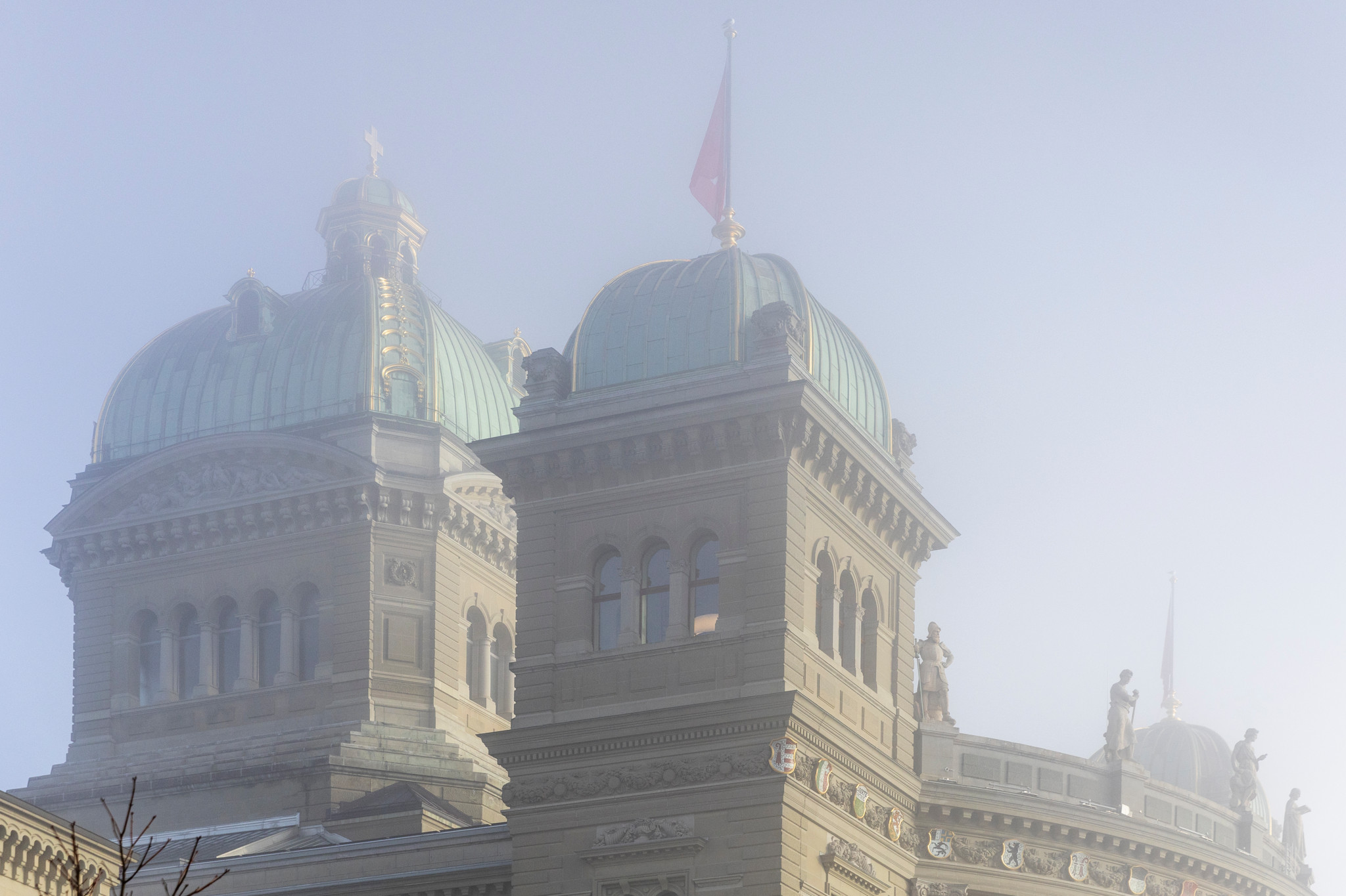 Kuppeln des Bundeshauses in Bern im dichten Nebel, mit einer Schweizer Fahne und Steinskulpturen an der Fassade.