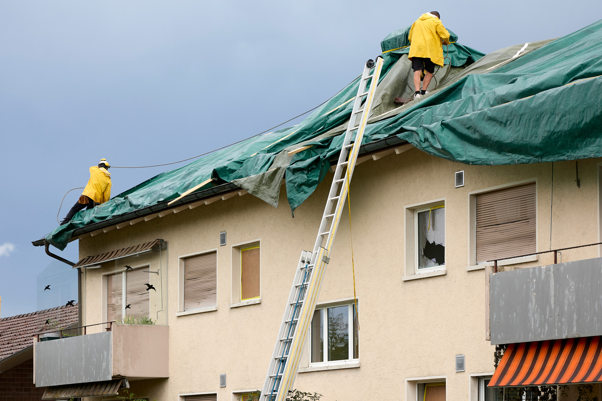 Zwei Arbeiter in gelben Jacken decken ein hagelzerstörtes Dach in Seuzach mit einer grünen Plane ab, erreichbar über eine Leiter.