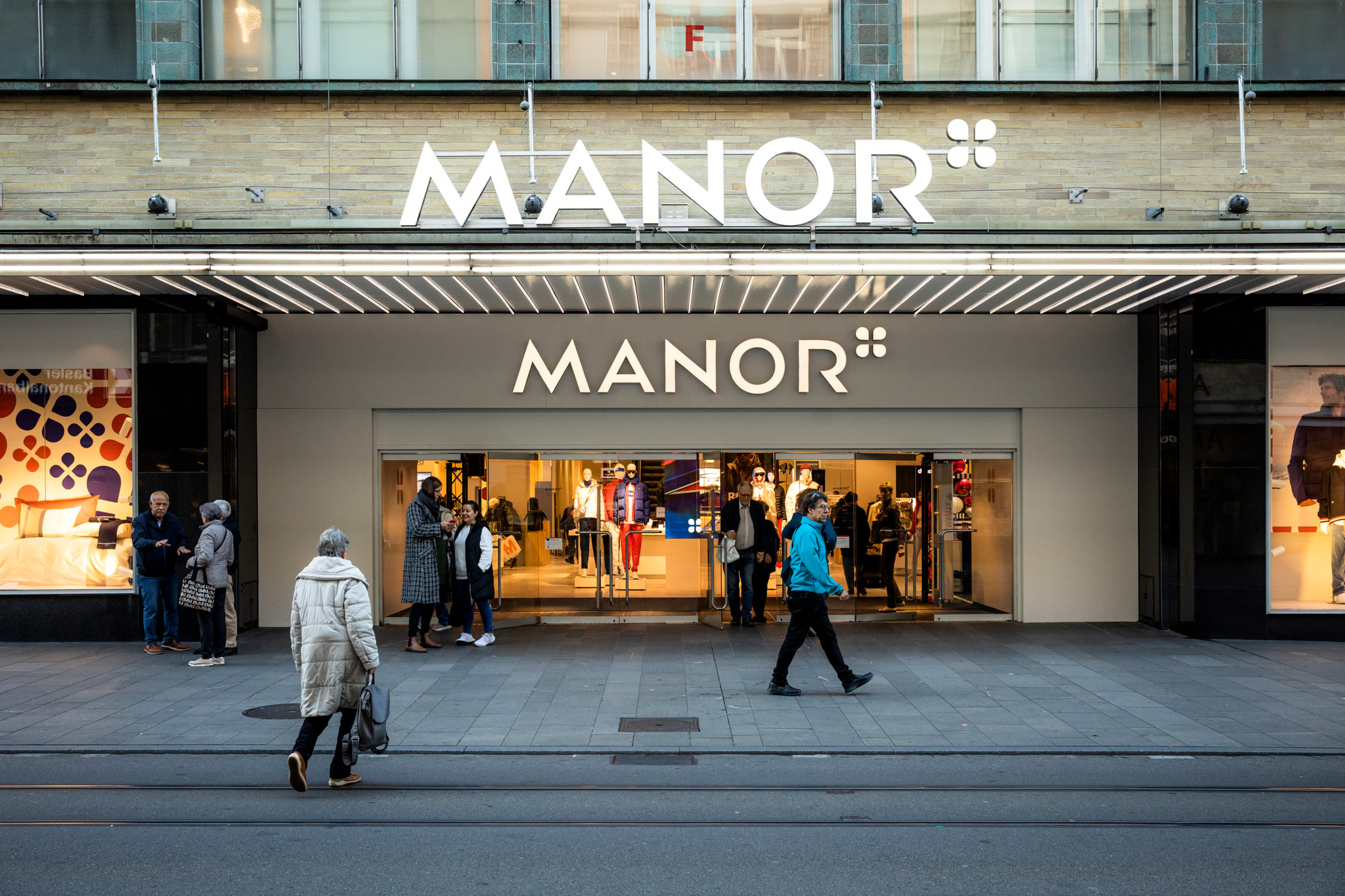 Eingangsbereich des Manor Basel mit neuem Logo, Menschen betreten und verlassen das Geschäft.