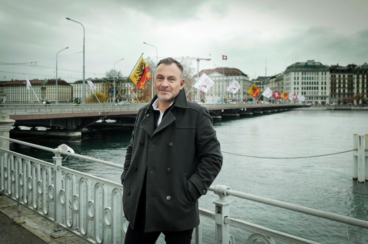 Pierre-Alain Dupraz, architecte, devant la passerelle piétonne de la rade à Genève, avec drapeaux en arrière-plan.