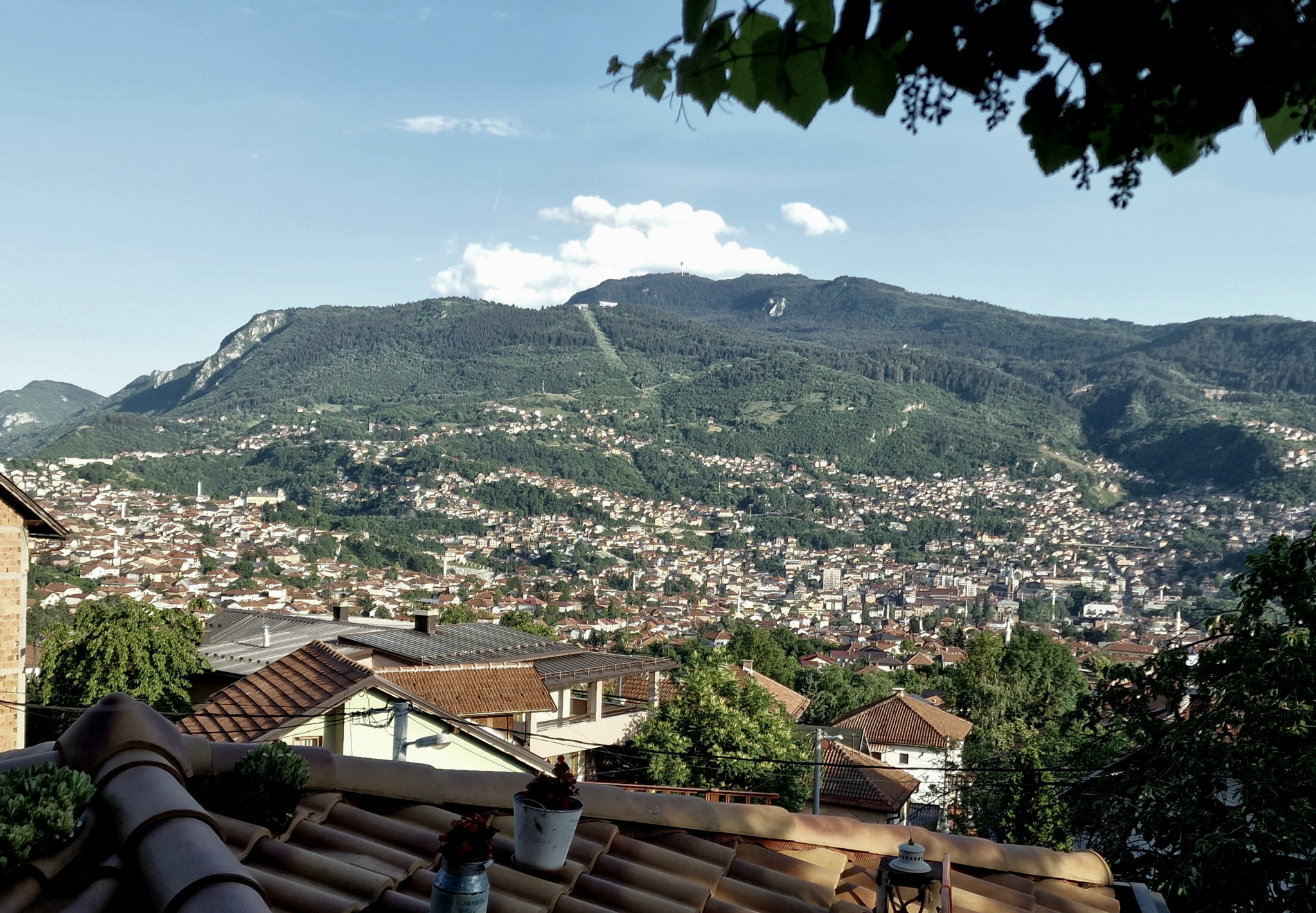 Blick auf eine Stadt in einem Tal, umgeben von bewaldeten Hügeln und Bergen, mit einem klaren blauen Himmel im Hintergrund.