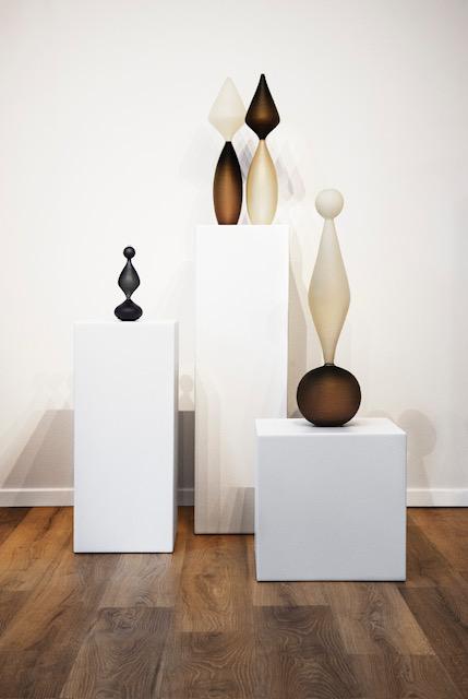 Sculptures en verre coloré sur des socles blancs dans une galerie d’art, posées sur un sol en bois.