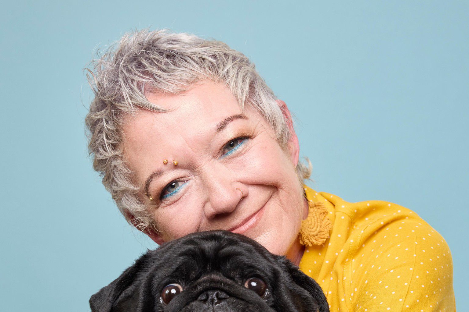 Femme souriante en chemise jaune tenant un carlin noir devant un fond bleu clair.