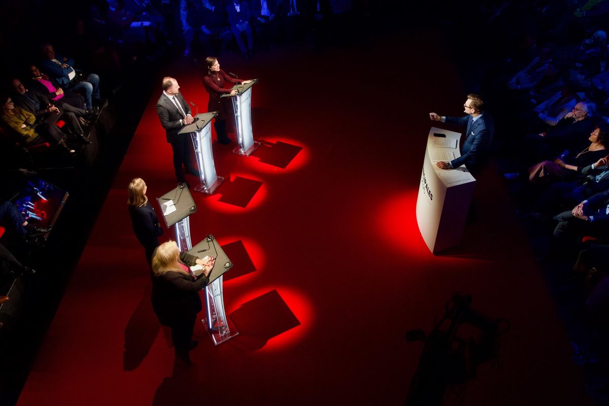 Débat politique avec quatre intervenants derrière des pupitres éclairés, face à un modérateur sur une scène rouge.