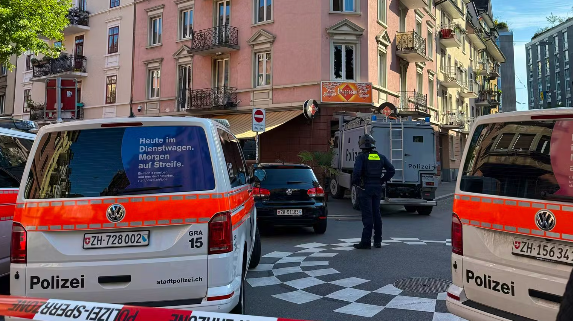 Polizeifahrzeuge und Beamte sperren eine Strassenecke in einer städtischen Umgebung mit Wohnhäusern ab.