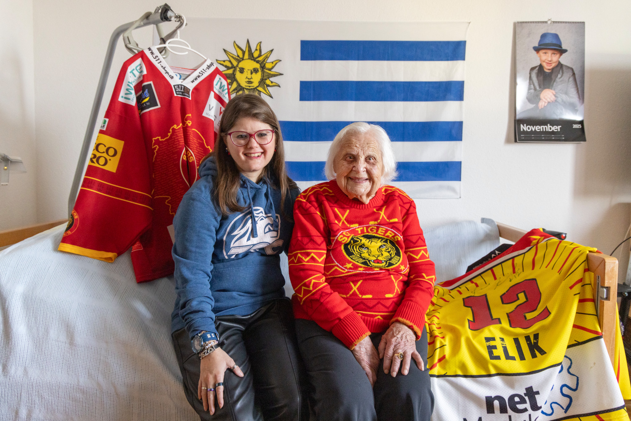 Anna Schmidhauser, eine fast 103-jährige Sportbegeisterte, sitzt mit ihrer Enkelin Lara Guggenbühl vor Trikots der SCL Tigers in einem Zimmer mit Uruguay-Flagge an der Wand.