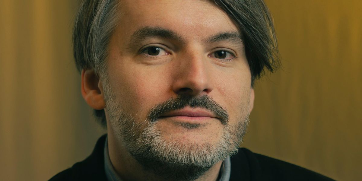 Saša Stanišić mit grauen Haaren und Bart, der ein schwarzes Oberteil vor einem gelben Hintergrund trägt.