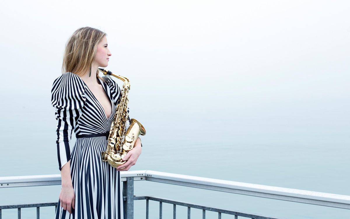 Femme en robe rayée tenant un saxophone sur une terrasse surplombant une mer calme.