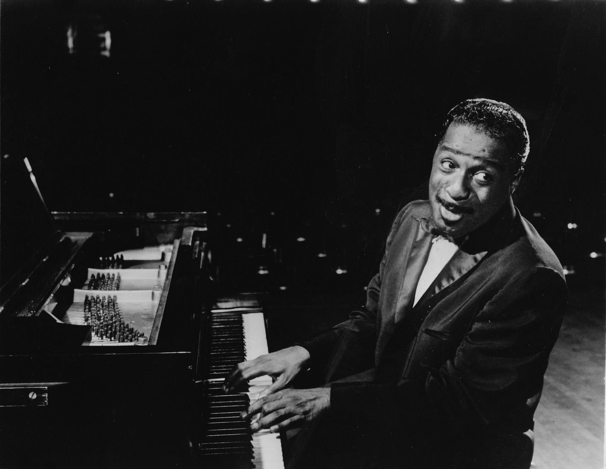 Erroll Garner joue du piano sur scène en 1965, souriant et élégant en costume sous un éclairage tamisé.