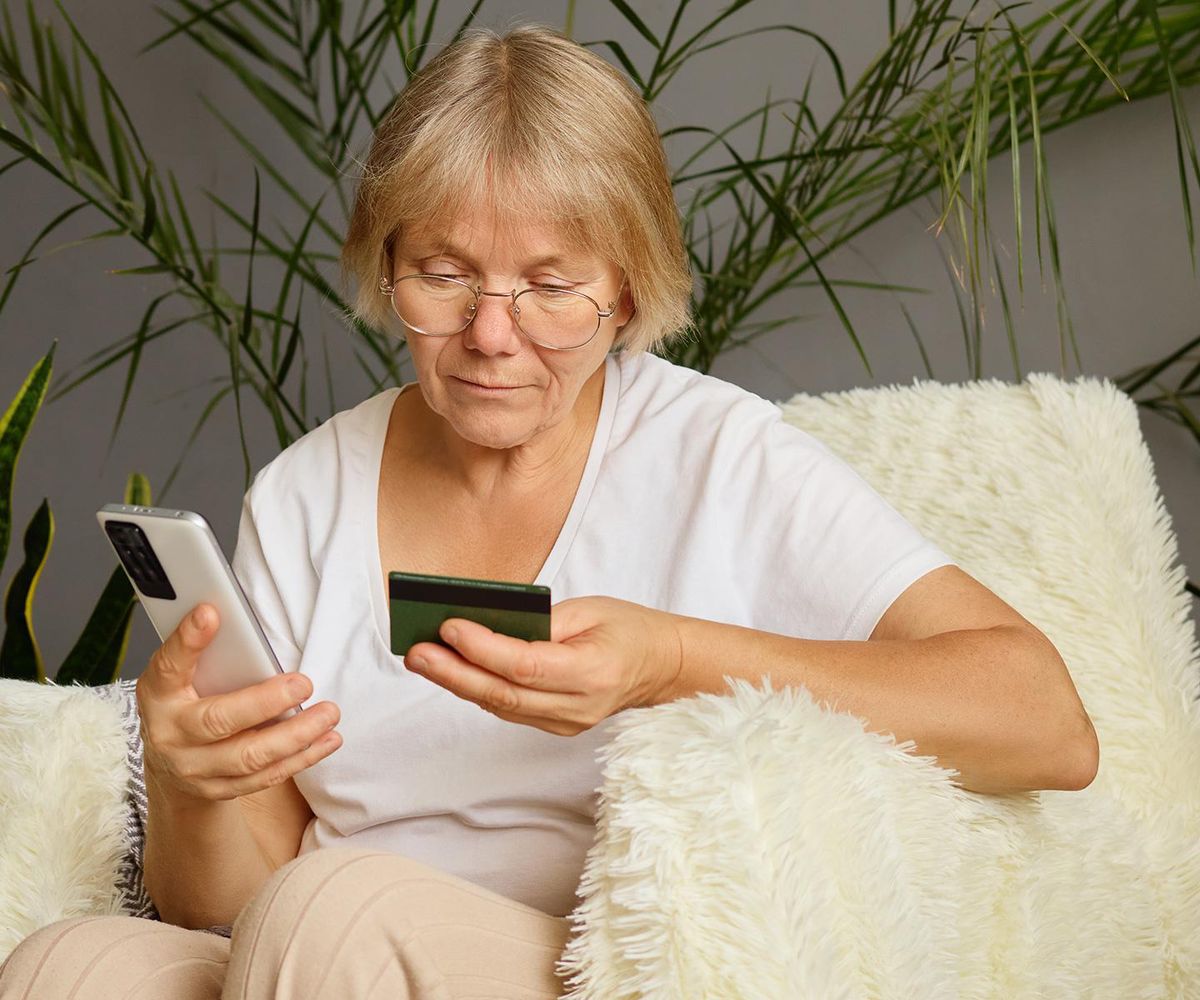 Une femme âgée avec lunettes tient un téléphone et une carte bancaire, assise sur un fauteuil moelleux.
