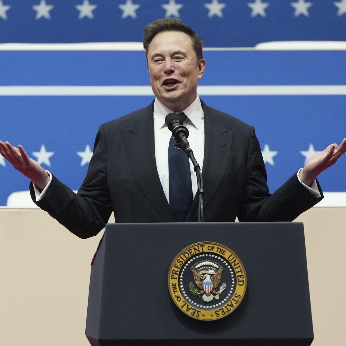 Homme en costume parlant devant un pupitre lors d’un événement présidential à Washington, avec un fond parsemé d’étoiles.