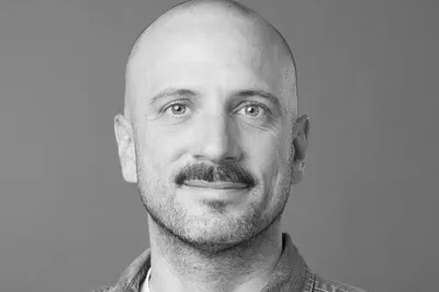 Portrait en noir et blanc d’un homme chauve avec une moustache, portant une chemise en jean et un t-shirt blanc.