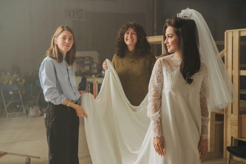 Une mariée en robe blanche et voile est entourée par deux femmes qui ajustent sa tenue dans un atelier de couture.