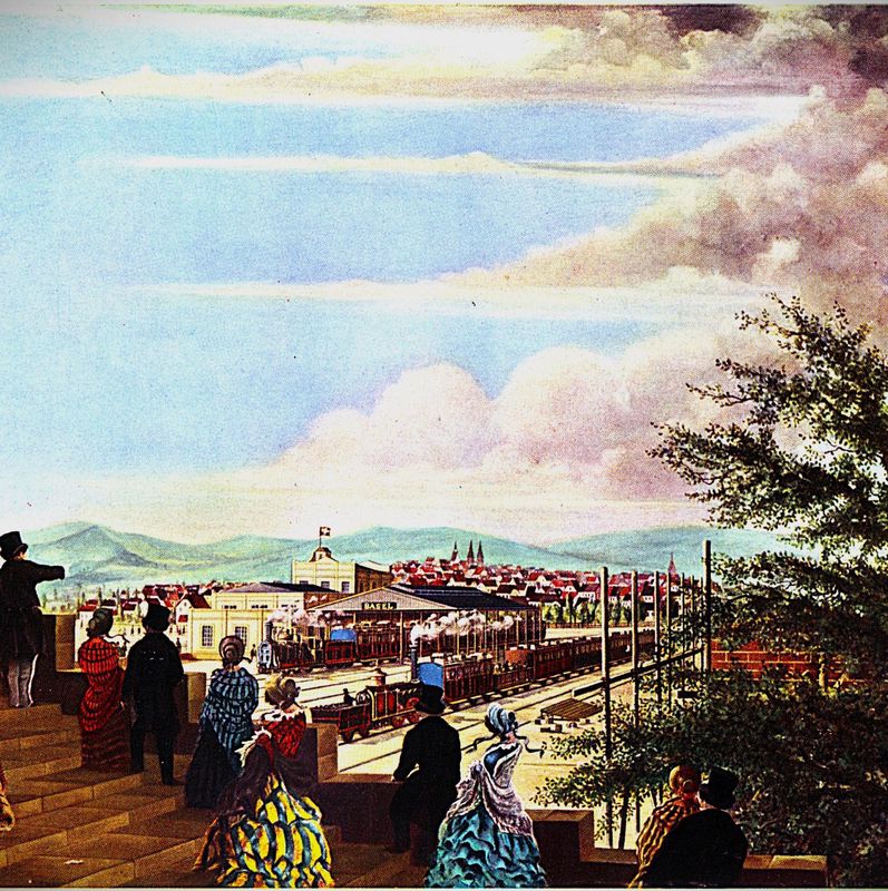 Historische Darstellung eines Bahnhofs im 19. Jahrhundert, mit versammelten elegant gekleideten Menschen. Eisenbahn und Gebäude im Hintergrund.