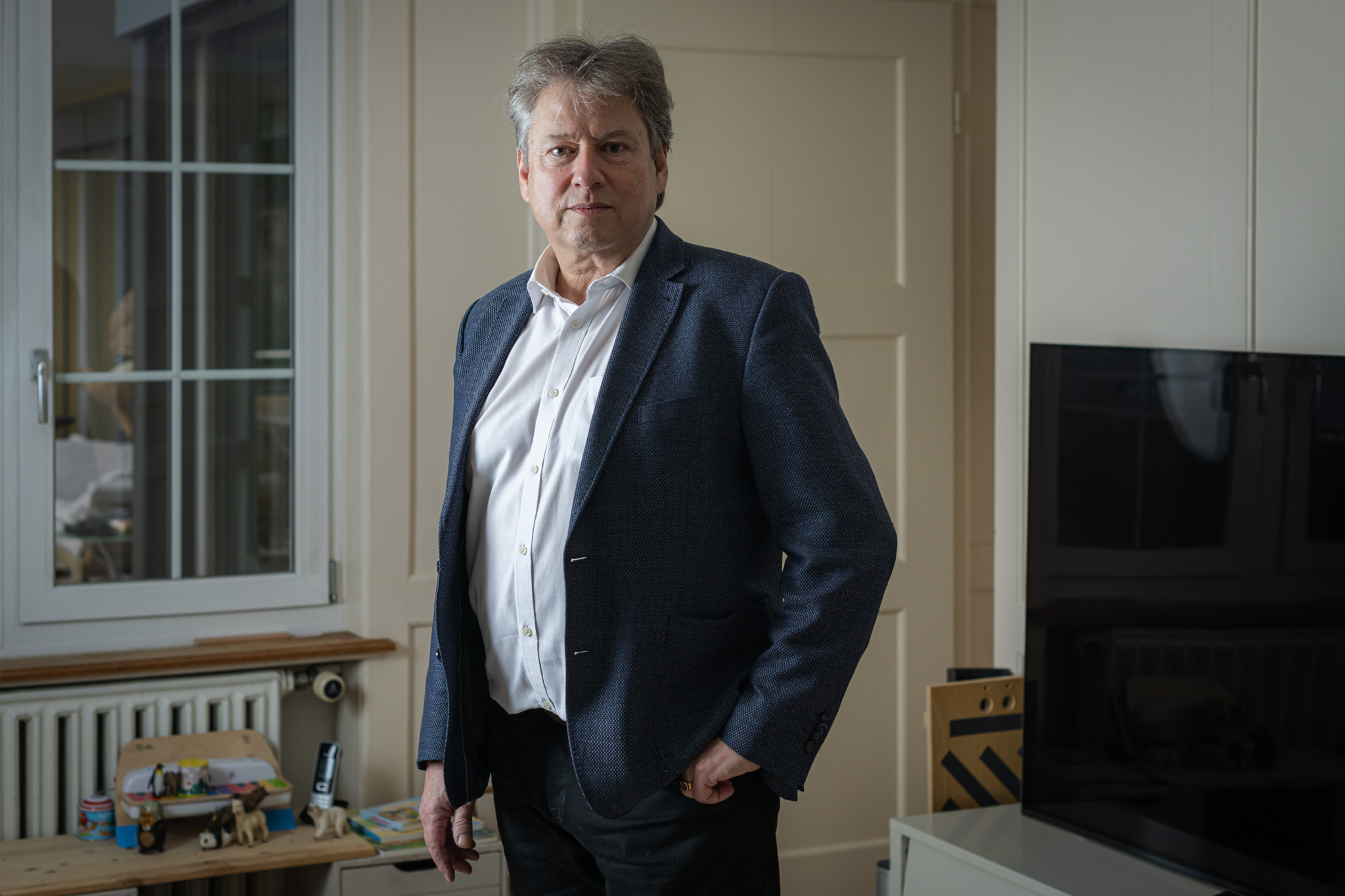 Reiner Eichenberger in seinem Zuhause in Herrliberg Feldmeilen, Portrait zur Halbierungsinitiative. Steht neben einem Fenster und einem Flachbildfernseher.