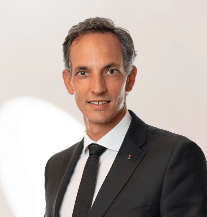 Homme en costume noir et cravate, avec un fond clair.