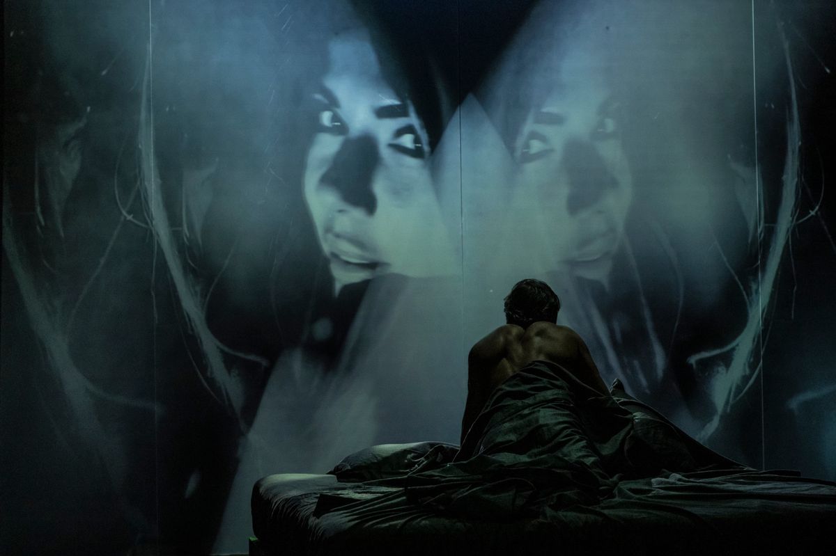 Un homme assis dans un lit sombre regardant un grand écran montrant des visages mystérieux.