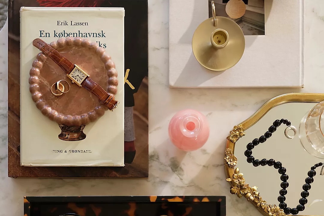 Stilvolle Anordnung auf einem Marmortisch mit einem Buch, einer Armbanduhr, goldenen Ringen und dekorativen Objekten, darunter eine Vase.