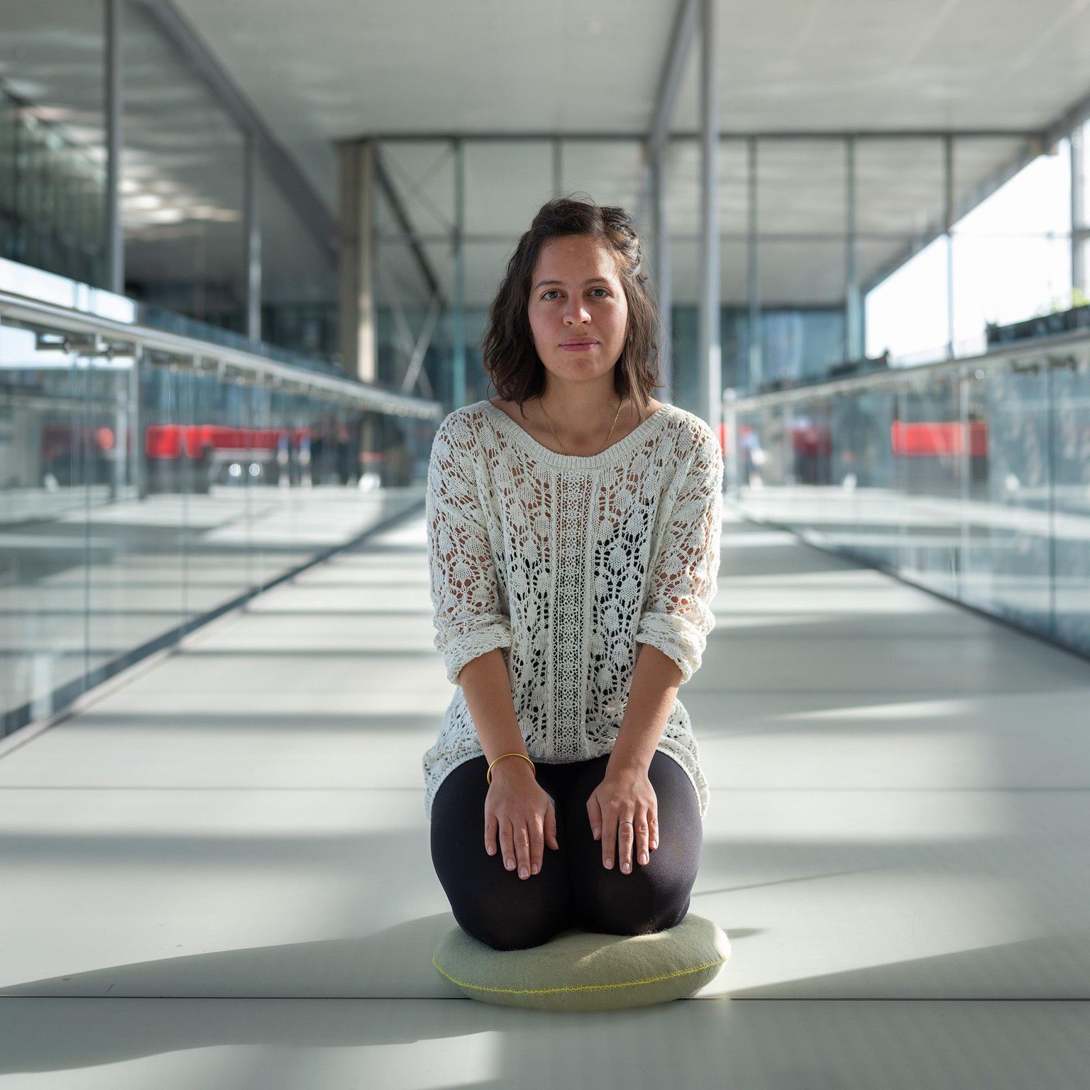 Une femme assise sur un coussin dans un espace moderne, lumineux et vaste avec de grandes fenêtres.